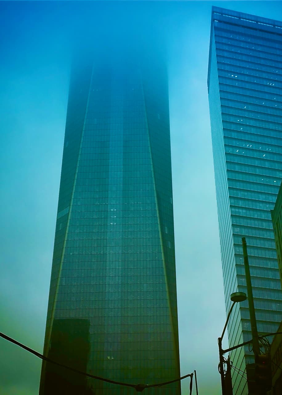 A tall glass tower disappearing into fog beside another high-rise.
