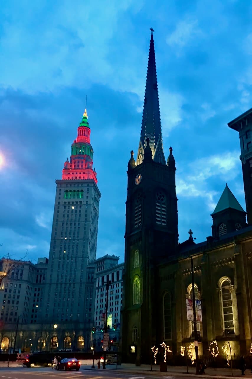 Downtown towers and a church spire at blue hour with colored lights in the skyline.