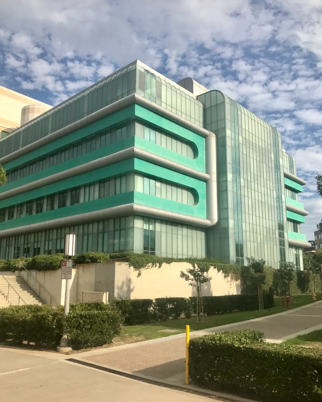An aqua and glass building with rounded forms, hedges, and a partly cloudy sky.