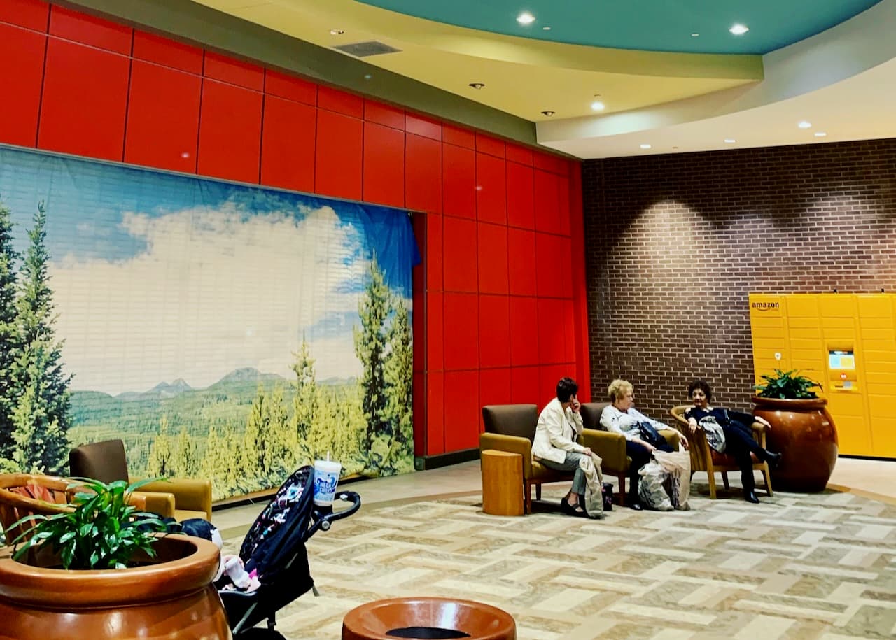 A colorful public lobby with red walls, yellow lockers, a scenic wall image, and people seated.