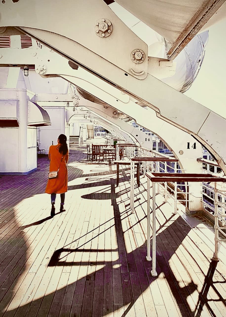 A person walking on a bright ship deck beneath curved white davits and long shadows.
