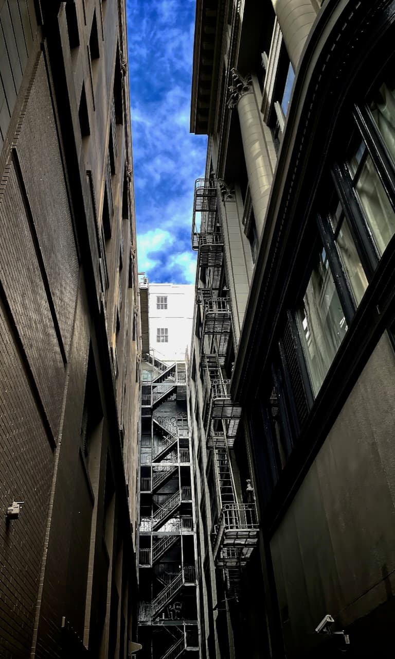 A city alley looking upward between tall buildings toward a bright blue sky.