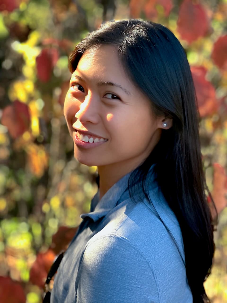 A close portrait of a smiling woman in front of softly blurred autumn leaves.