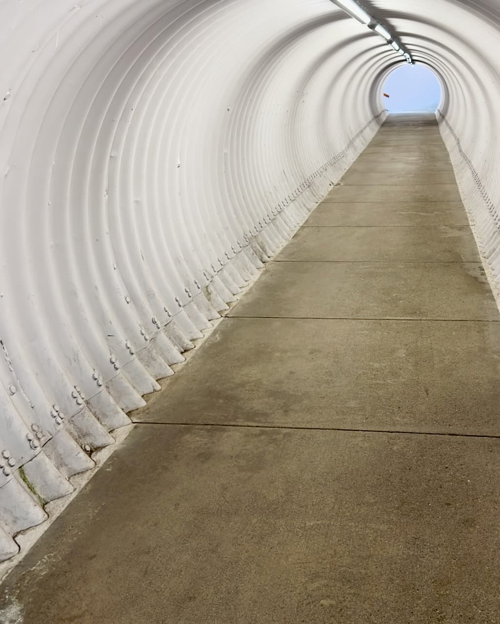 A white ribbed pedestrian tunnel with a concrete path rising toward a circular opening.