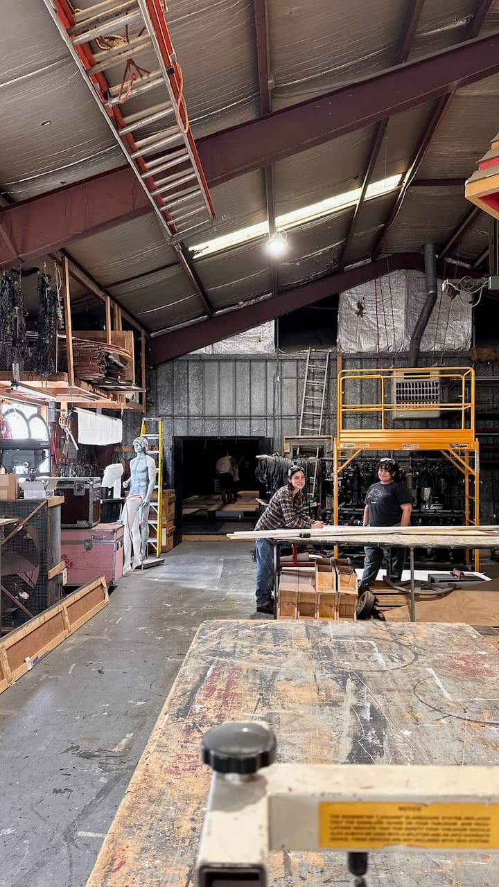 A scene shop interior with ladders, platforms, lumber, and people working at a table.