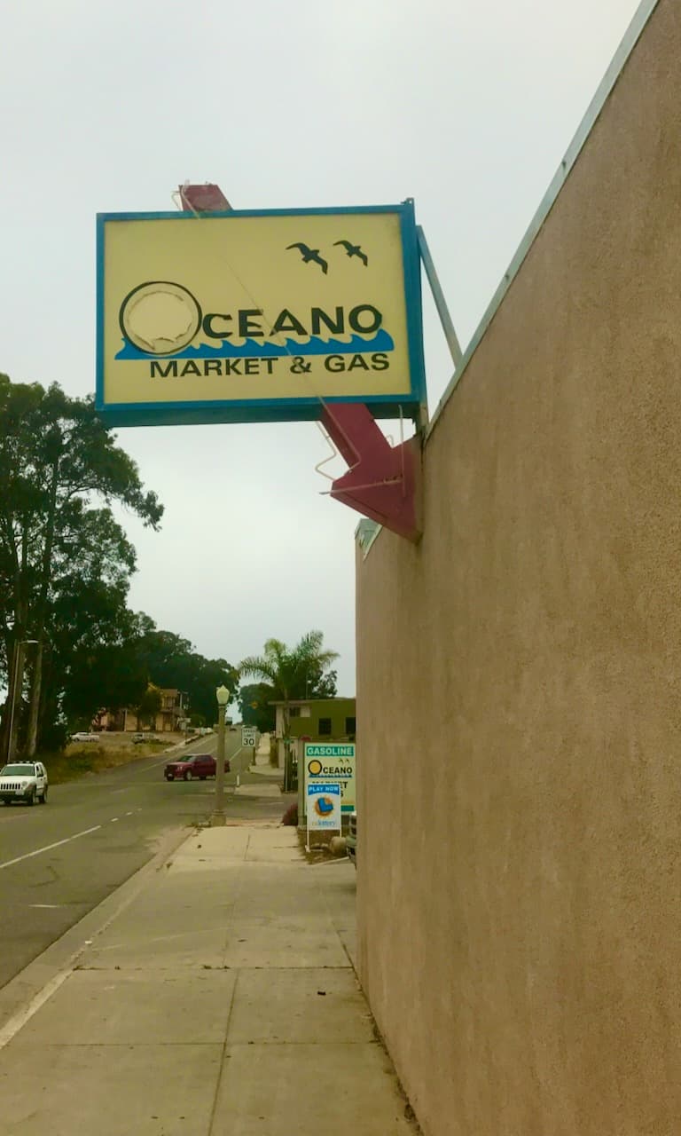 An Oceano Market and Gas sign mounted above a sidewalk and roadside wall.