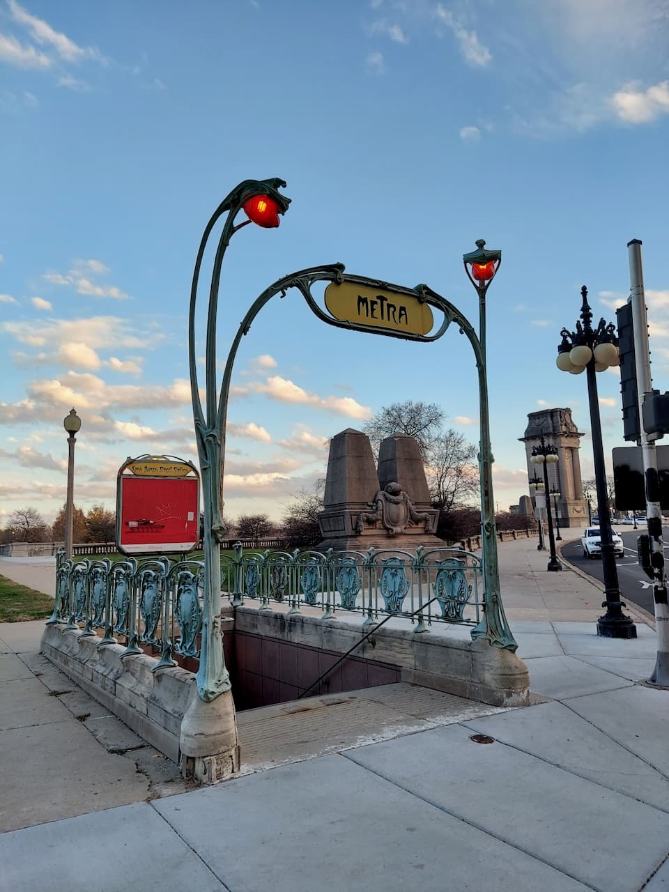 A Metra entrance with ornate metalwork, red lamps, and a blue evening sky.