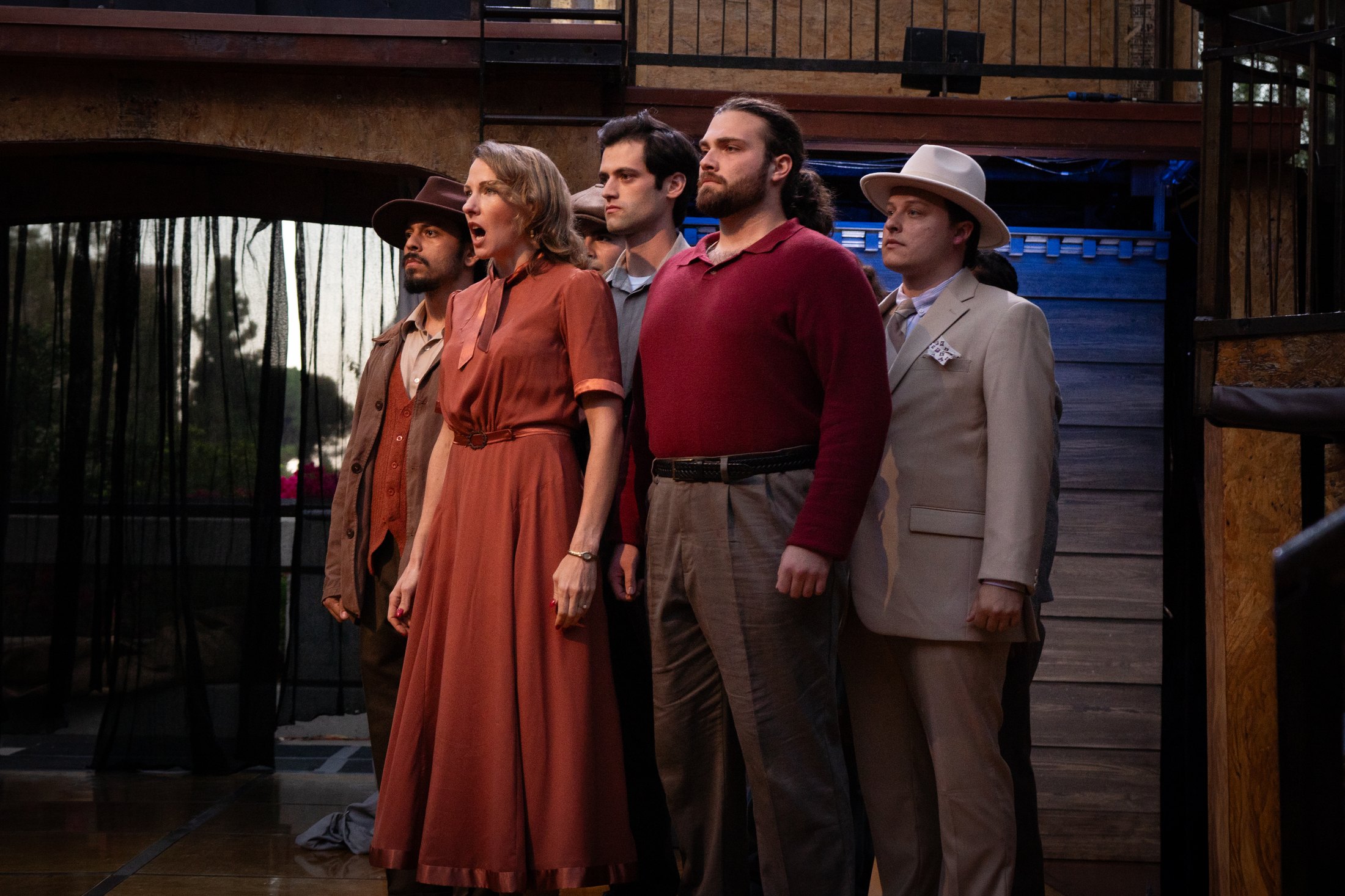 The ensemble gathers in earth-toned costumes before the scenic structure.