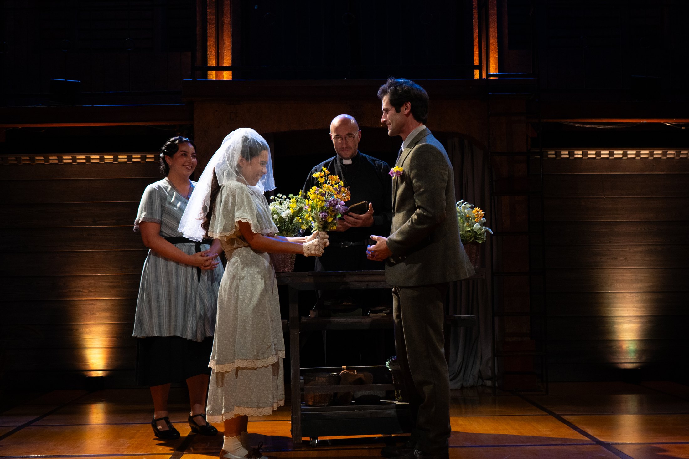 The company gathers around a flower-covered table within the Depression-era setting.