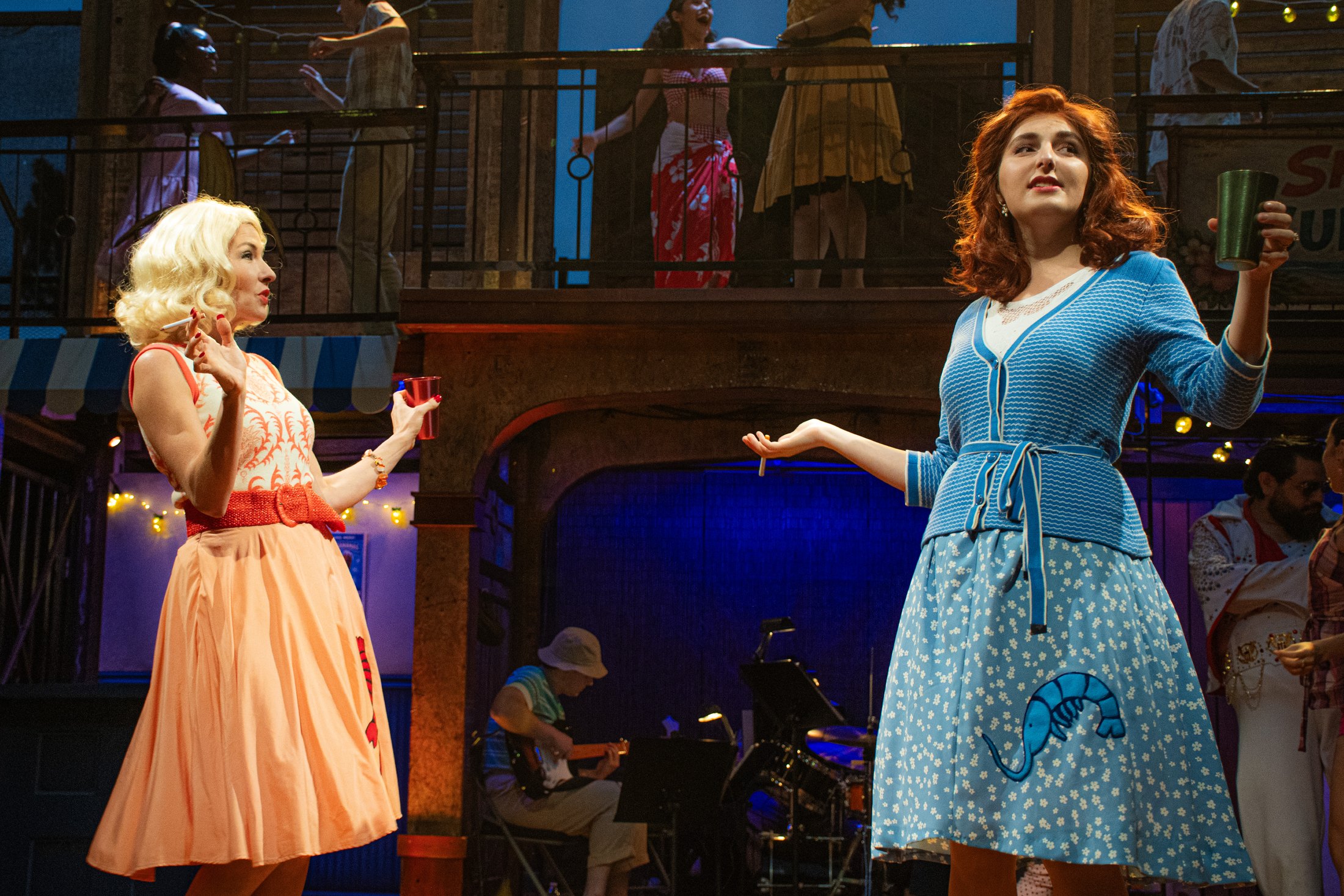 The merry wives share a scene against the layered mid-century beach-town architecture.