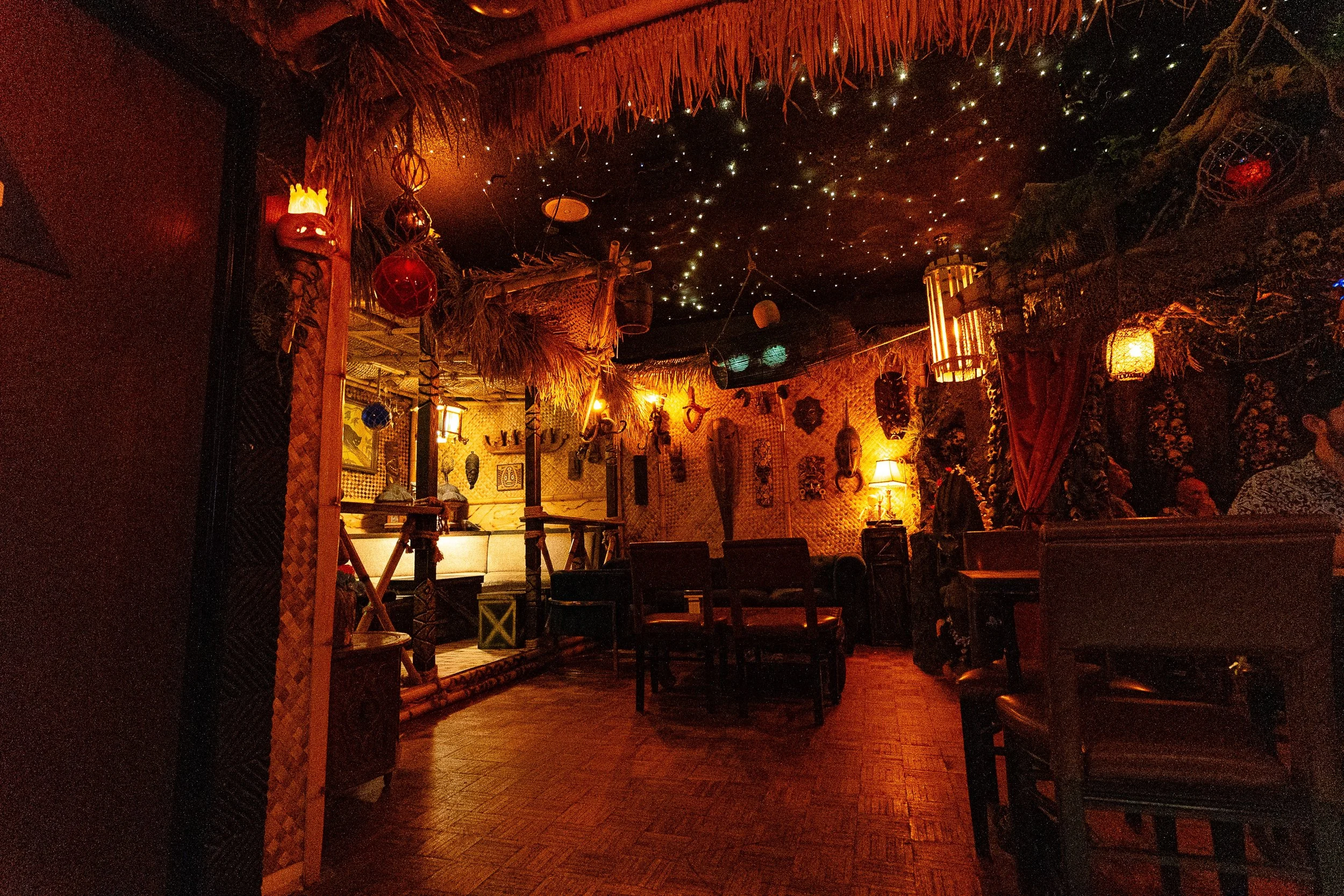 Lounge seating inside STOWAWAY with thatched overhead details, warm amber light, masks, and a starry ceiling.